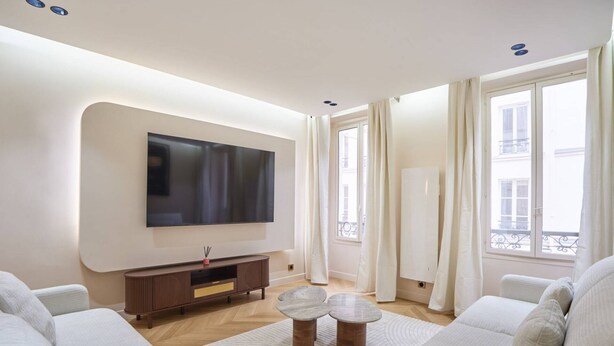 A modern living room with a flat-screen TV, a wooden entertainment unit, two round coffee tables, and a large window with curtains. - Caire 3 - One Bedroom Apartment, Sleeps 6 (Paris)