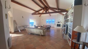 Amplia sala de estar con chimenea, un sofá y una mesa de comedor. Hay un balcón con vistas al mar.