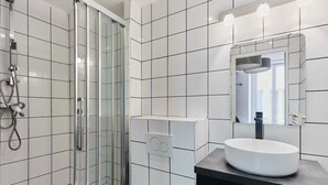 A modern bathroom with white tiled walls, a glass shower enclosure, a white sink with a dark countertop, and a mirror with lights above it. - Caire 5 - Studio Apartment, Sleeps 2 (Paris)
