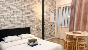 A bedroom with a brick wall, a bed with white bedding, a small table with chairs, and a wardrobe. - Charon 2 - Studio Apartment, Sleeps 2 (Paris)