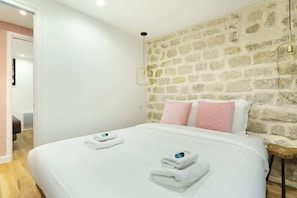 Une chambre avec un mur en pierre, un lit avec une literie blanche et des oreillers roses, et deux serviettes posées sur le lit.