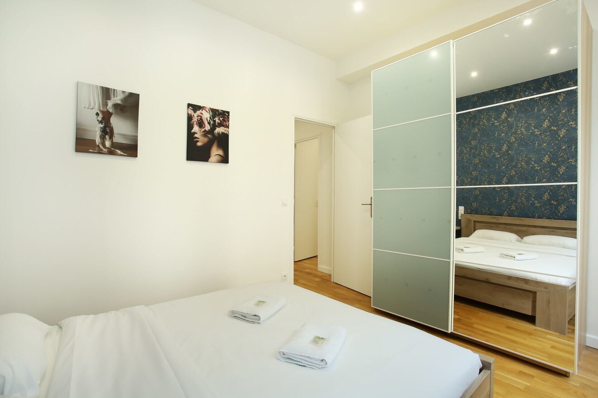 A bedroom with a bed, two towels, a wardrobe with a mirror, and two framed pictures on the wall.
