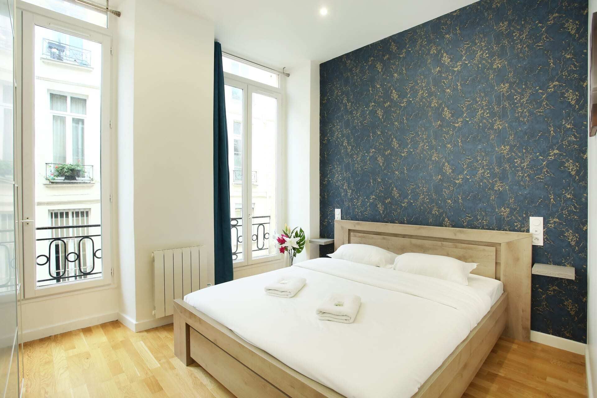 A bedroom with a large bed, a window with a view of buildings, a radiator, and a dark blue wall with a floral pattern.