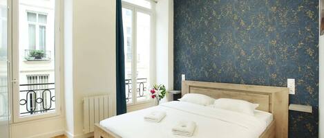 A bedroom with a large bed, a window with a view of buildings, a radiator, and a dark blue wall with a floral pattern.