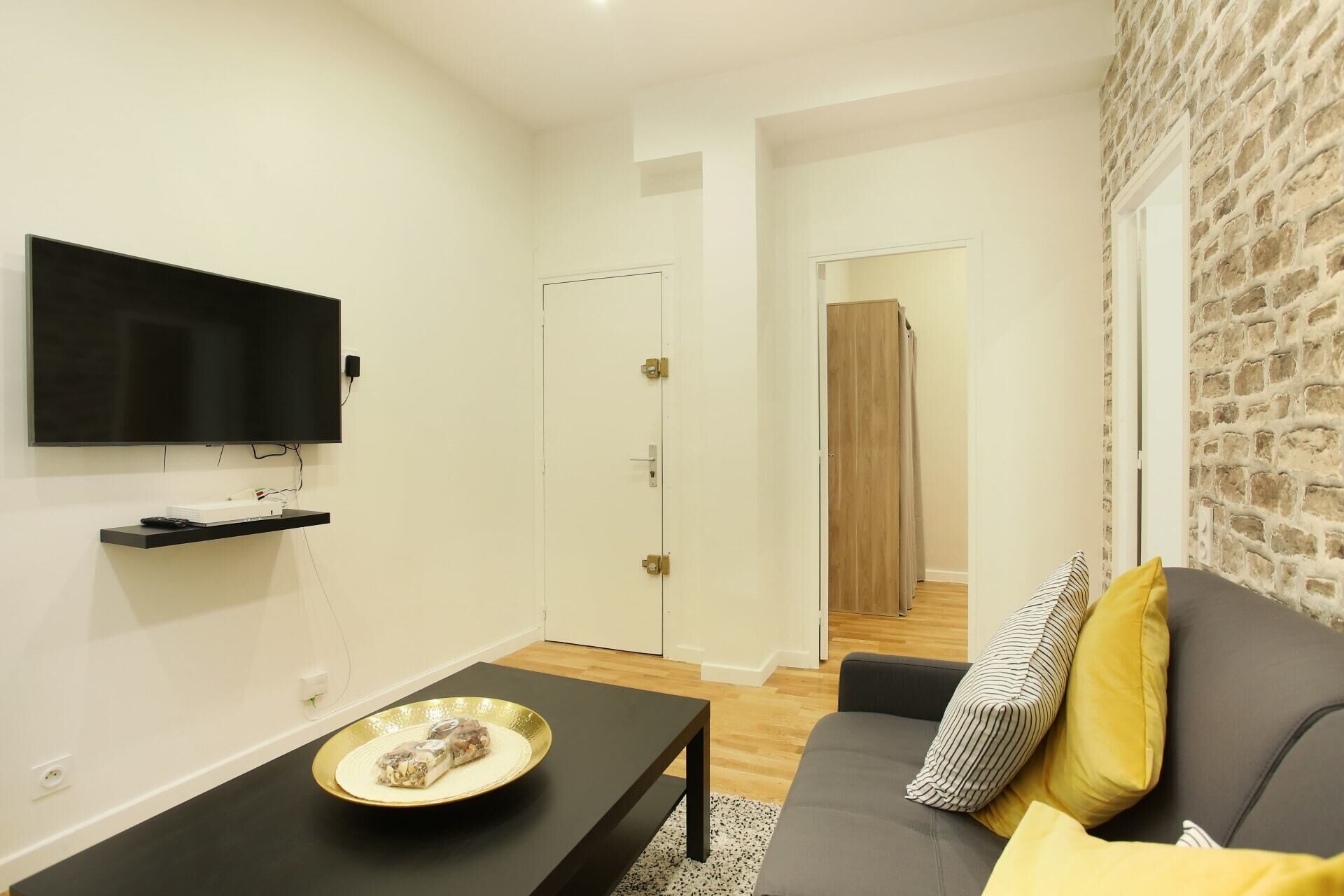 Una sala de estar moderna con un televisor de pantalla plana, una mesa de centro negra con un plato decorativo, un sofá gris con cojines amarillos y una pared con textura de piedra.