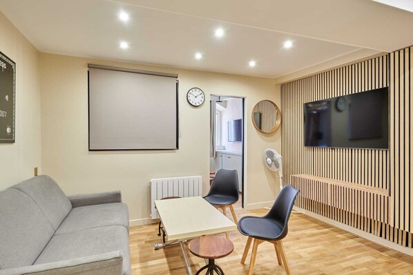 A living room with a sofa, a table, two chairs, a TV, a clock, and a radiator.