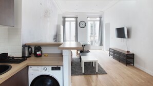 A compact kitchen with a washing machine, a dining area with a table and chairs, a TV mounted on the wall, and large windows.