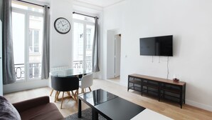 A modern living room with a glass dining table, black and white chairs, a black coffee table, a brown sofa, a TV mounted on the wall, and a large window with curtains.