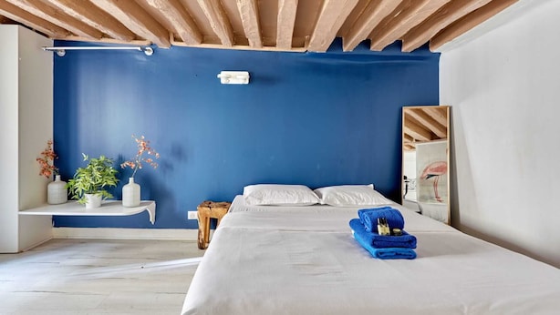 Un dormitorio con pared azul, vigas de madera en el techo, una cama con ropa blanca, una toalla azul y una mesita con una planta.