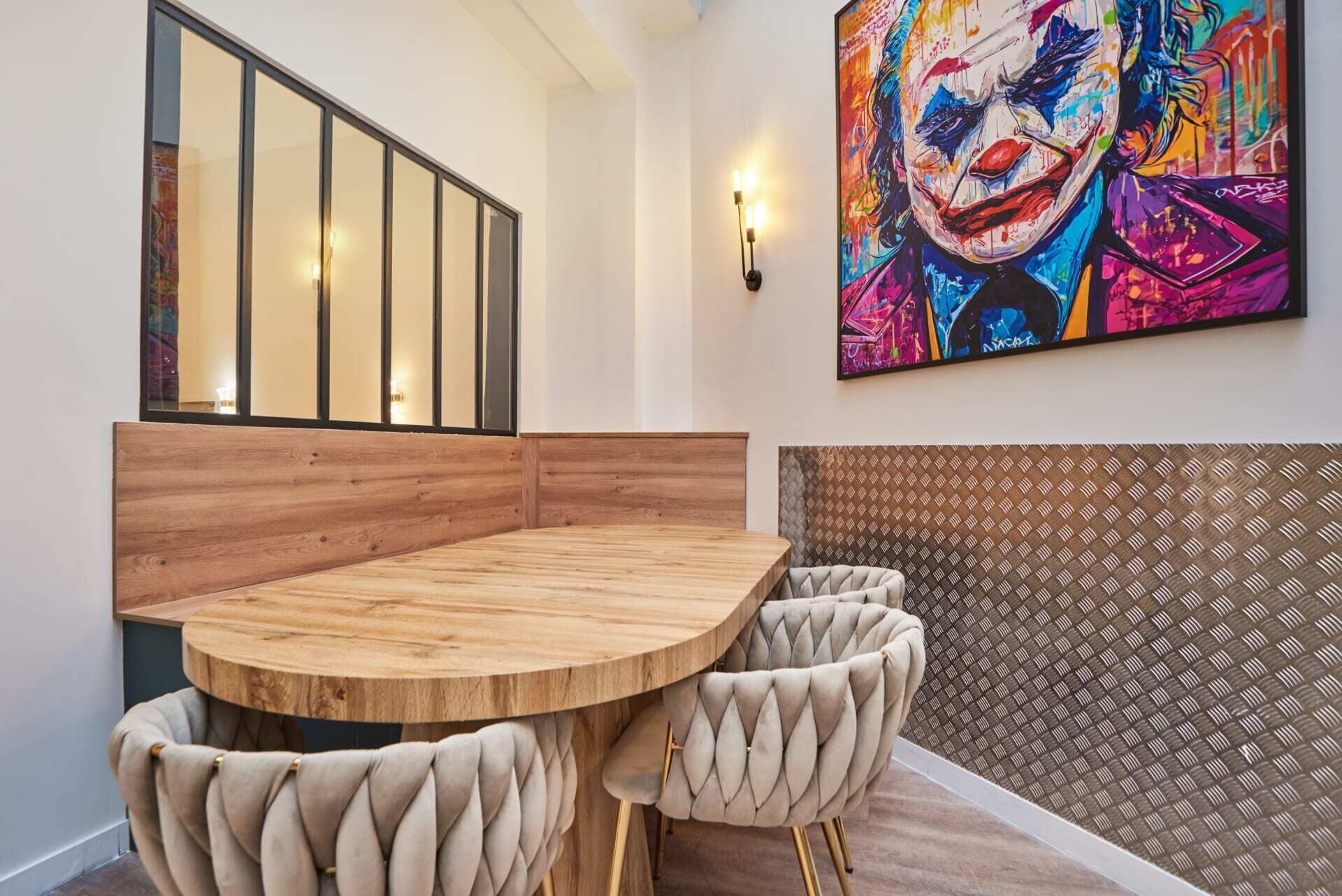 Un coin repas avec une table ronde en bois et des chaises à motifs capitonnés, un grand tableau coloré au mur et un mur à motifs derrière les sièges.