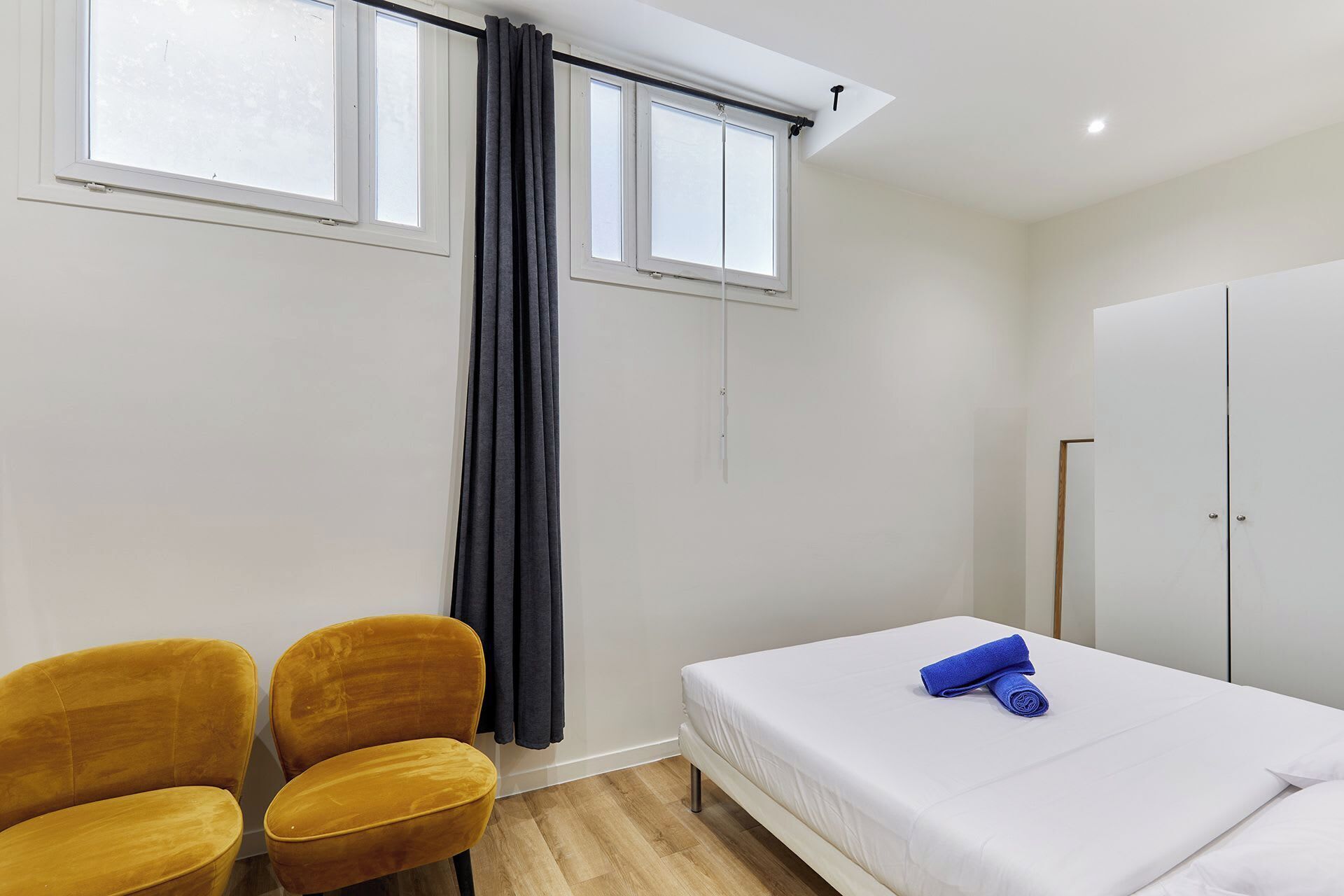 A bedroom with a bed, two chairs, a wardrobe, and a window with curtains.