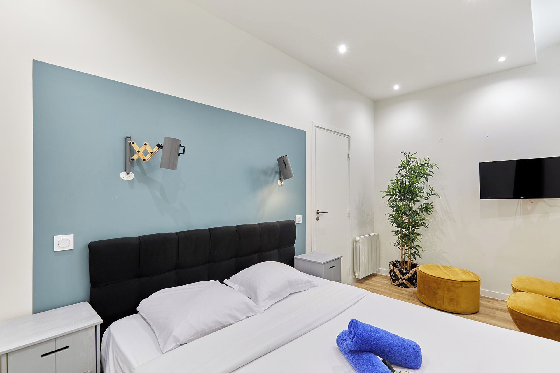 Een moderne hotelkamer met een bed, nachtkastjes, een aan de muur bevestigde televisie, een bloempot en een klein krukje.