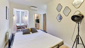Une chambre avec un lit, un bureau et une chaise. On aperçoit un coin cuisine au fond.