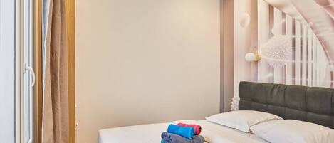 Una cama bien hecha con almohadas y toallas dobladas en una habitación con piso de madera y pared blanca.