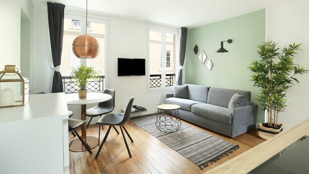 A modern living room with a grey sofa, a round coffee table, and a dining area with a white table and chairs. There is a large window with curtains, a flat-screen TV mounted on the wall, and a potted plant. - Meslay 9 - One Bedroom Apartment, Sleeps 4 (Paris)