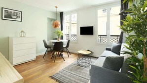 A modern living room with a grey sofa, a dining table with chairs, a TV mounted on the wall, and a large window with a view of the outside. - Meslay 9 - One Bedroom Apartment, Sleeps 4 (Paris)