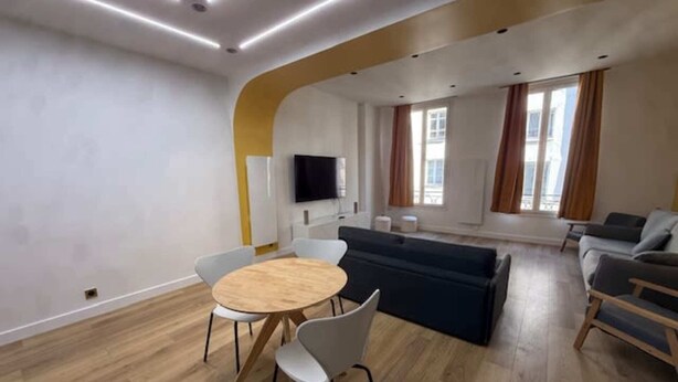 A modern living room with a round wooden table, white chairs, a dark sofa, and a television mounted on the wall. - St Denis 2 - One Bedroom Apartment, Sleeps 6 (Paris)