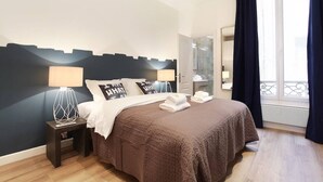 Une chambre d’hôtel avec un lit, deux lampes de chevet, une table de nuit, une salle de bain visible à travers une porte ouverte et un balcon avec des rideaux.