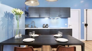 Une cuisine moderne avec des meubles de couleur sombre, un plan de travail blanc et un coin repas agrémenté d’une table en bois mise pour deux personnes.