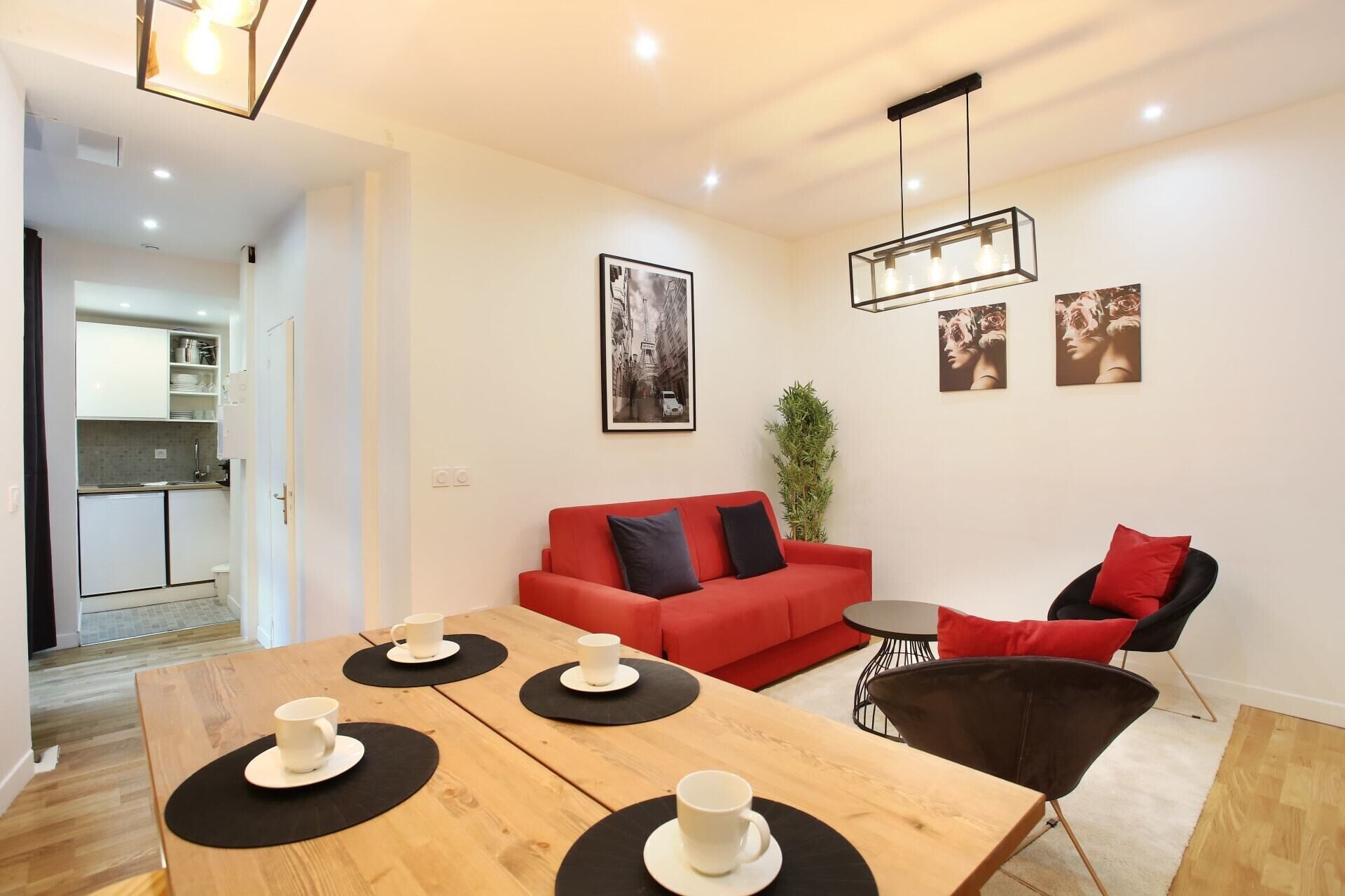 Un salon moderne avec un canapé rouge, une table à manger en bois sur laquelle sont posées quatre tasses blanches, une chaise noire et une petite table d’appoint.