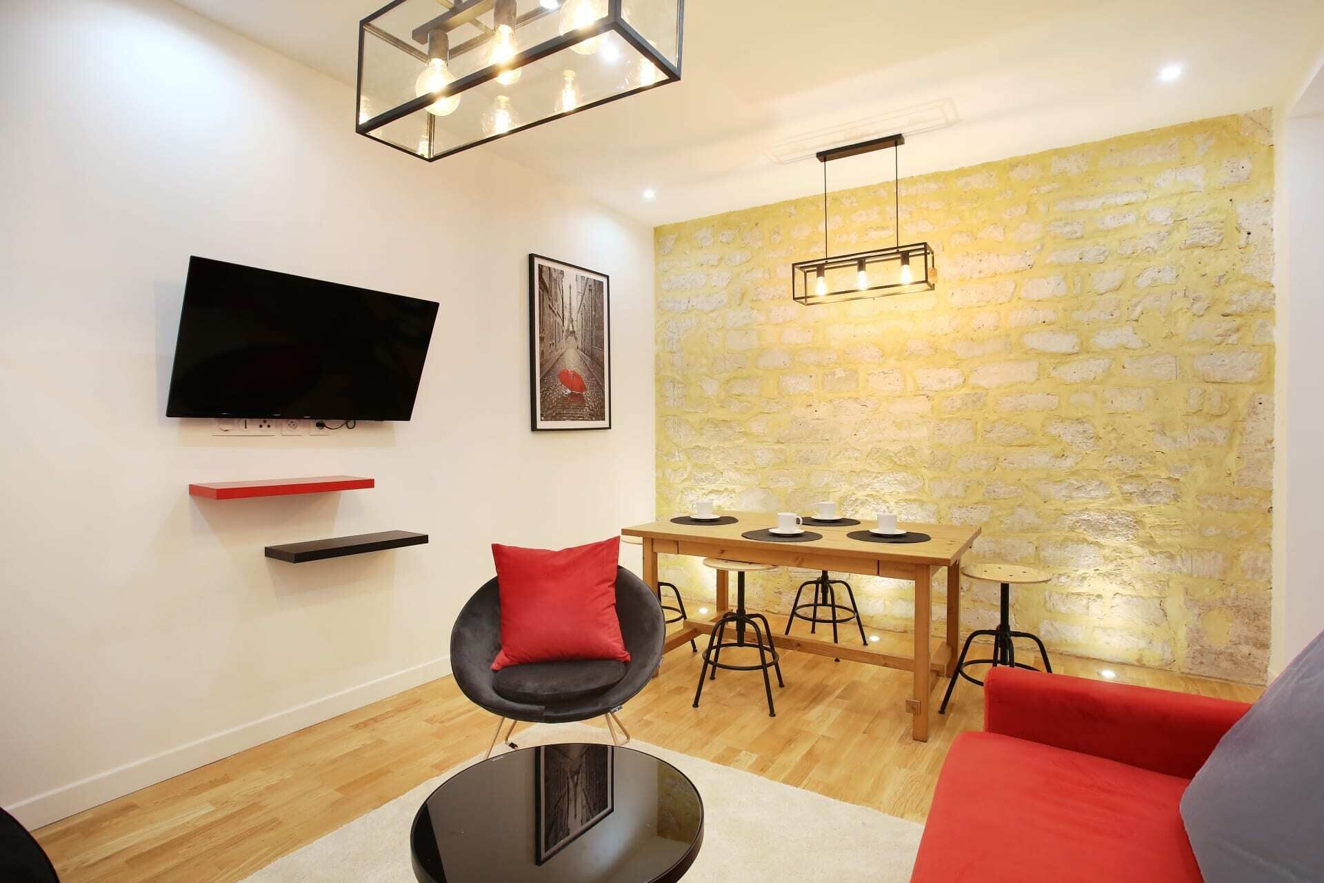 A modern living room with a flat-screen TV, a dining table with stools, a red sofa, and a stone wall.