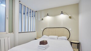 A bedroom with a large bed, two bedside tables, a window with blinds, and a wall-mounted lamp.