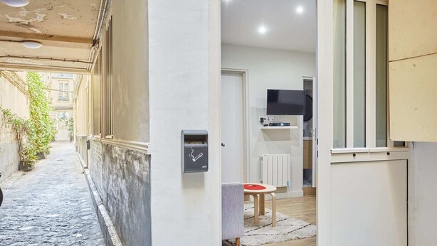 A narrow alleyway with a stone floor, a white wall with a mailbox, and a door leading to a room with a television and a small table.