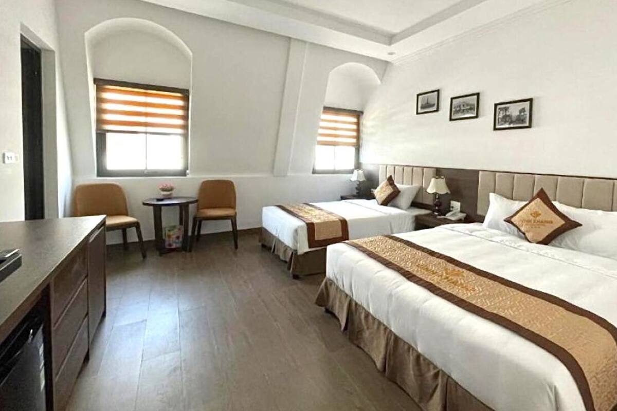 A hotel room with two beds, a desk, and two chairs. There are two windows with blinds, and the walls are decorated with framed pictures.