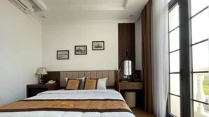 A hotel room with a large bed, a bedside table with a lamp, a desk with a chair, and a large window with curtains. - ViNH KHANG HA LONG HOTEL (Bai Chay)