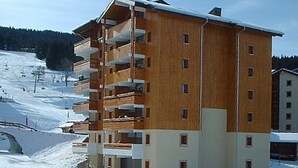 Un edificio de madera de varios pisos con balcones, ubicado en una zona nevada con una pista de esquí al fondo.