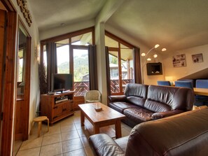 Ein Wohnzimmer mit einer Couch, einem Fernseher, einem Holzschrank und einem Balkon mit Blick ins Freie.