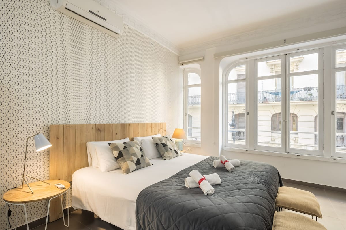 Hotellihuone, jossa on suuri sänky, puinen pääty, pyöreä yöpöytä, lamppu ja suuret ikkunat, joista avautuu näkymä kaupunkiin.