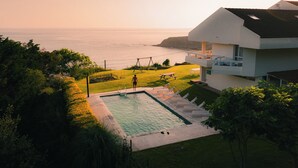 Piscina con vista al océano, rodeada por un césped cuidado y una casa moderna.