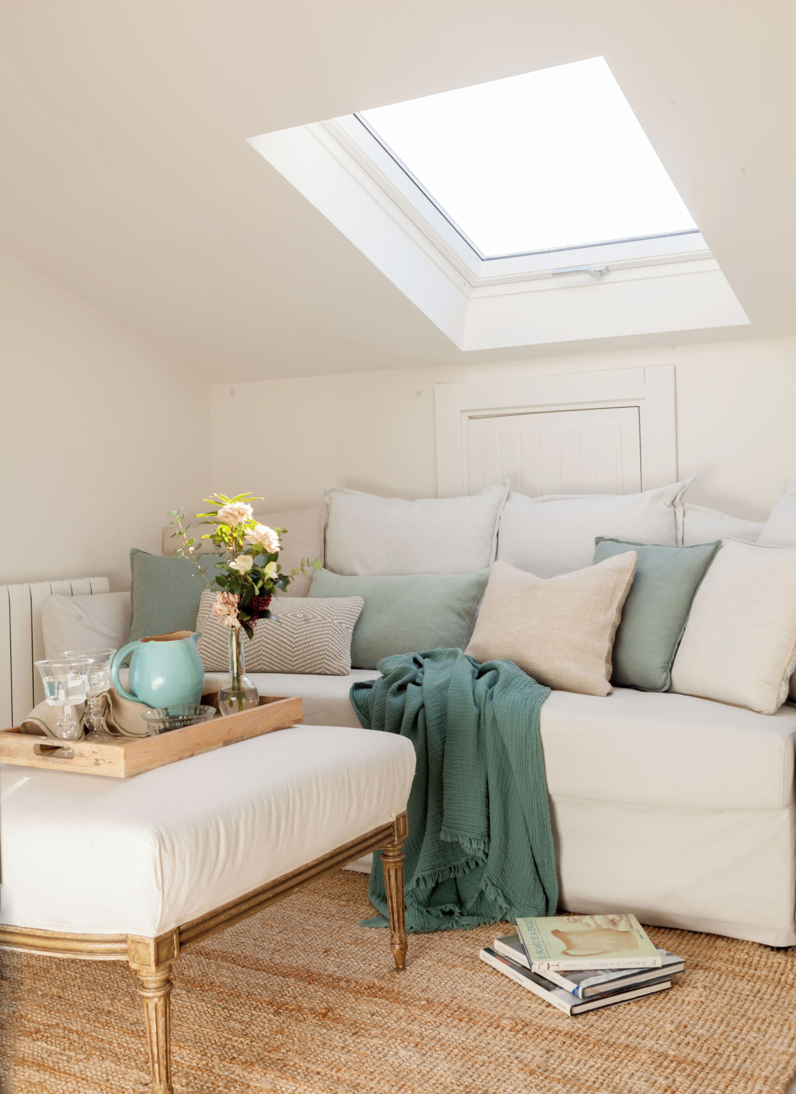 A cozy room with a skylight, a wooden bench, a tray with glasses and a pitcher, a green blanket, and a stack of books on the floor.