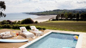 A pool with a view of the beach and hills, surrounded by lounge chairs and a wooden deck. - Wishome—A special villa to extend your summer at Oyambre Beach, Comillas (San Vicente de la Barquera)