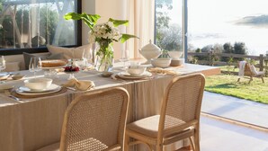 A dining area with a table set for two, featuring wicker chairs, a vase with flowers, and a view of a lake through large windows. - Wishome—A special villa to extend your summer at Oyambre Beach, Comillas (San Vicente de la Barquera)