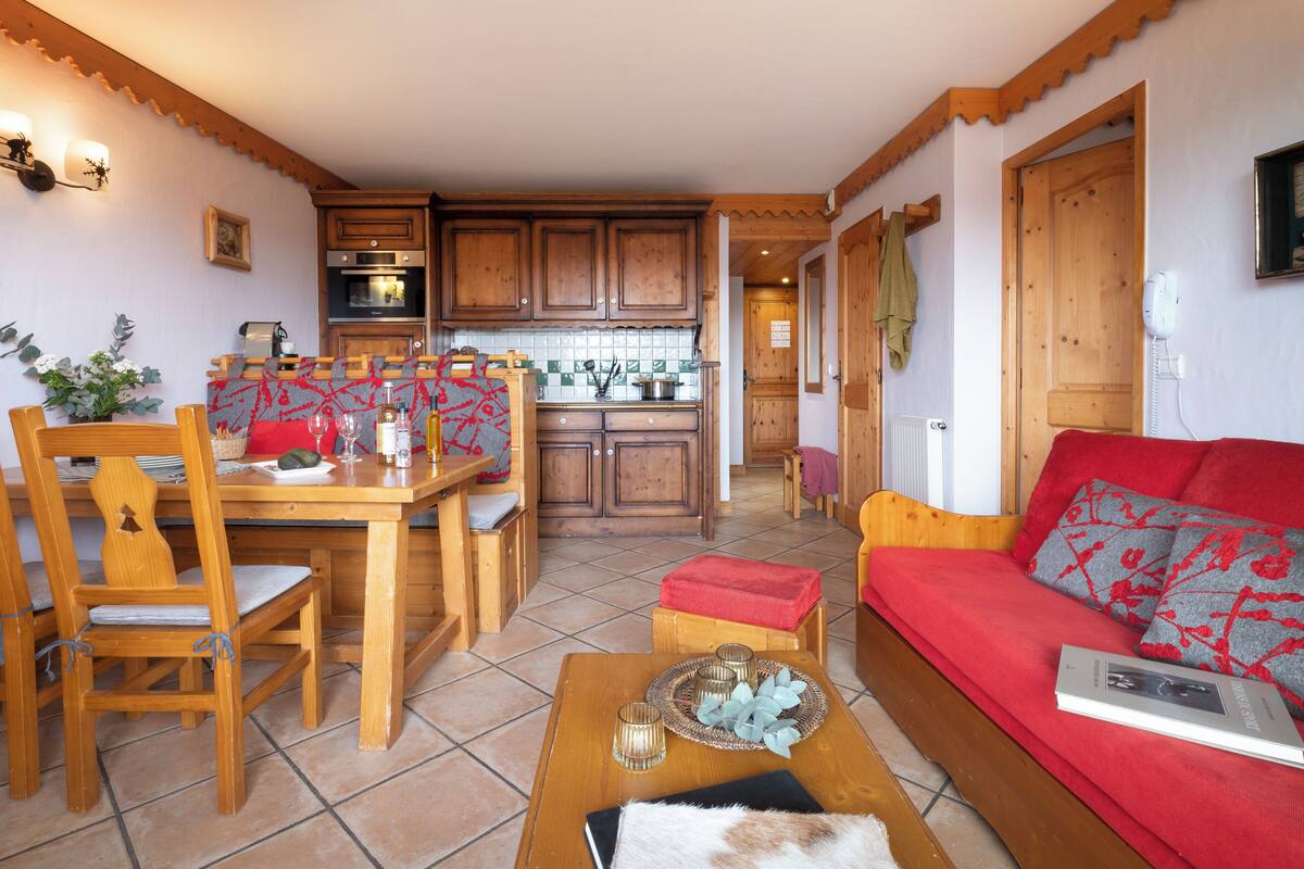 A cozy living room with a red sofa, wooden dining table, and chairs. A kitchen with wooden cabinets and a tiled backsplash is visible in the background.