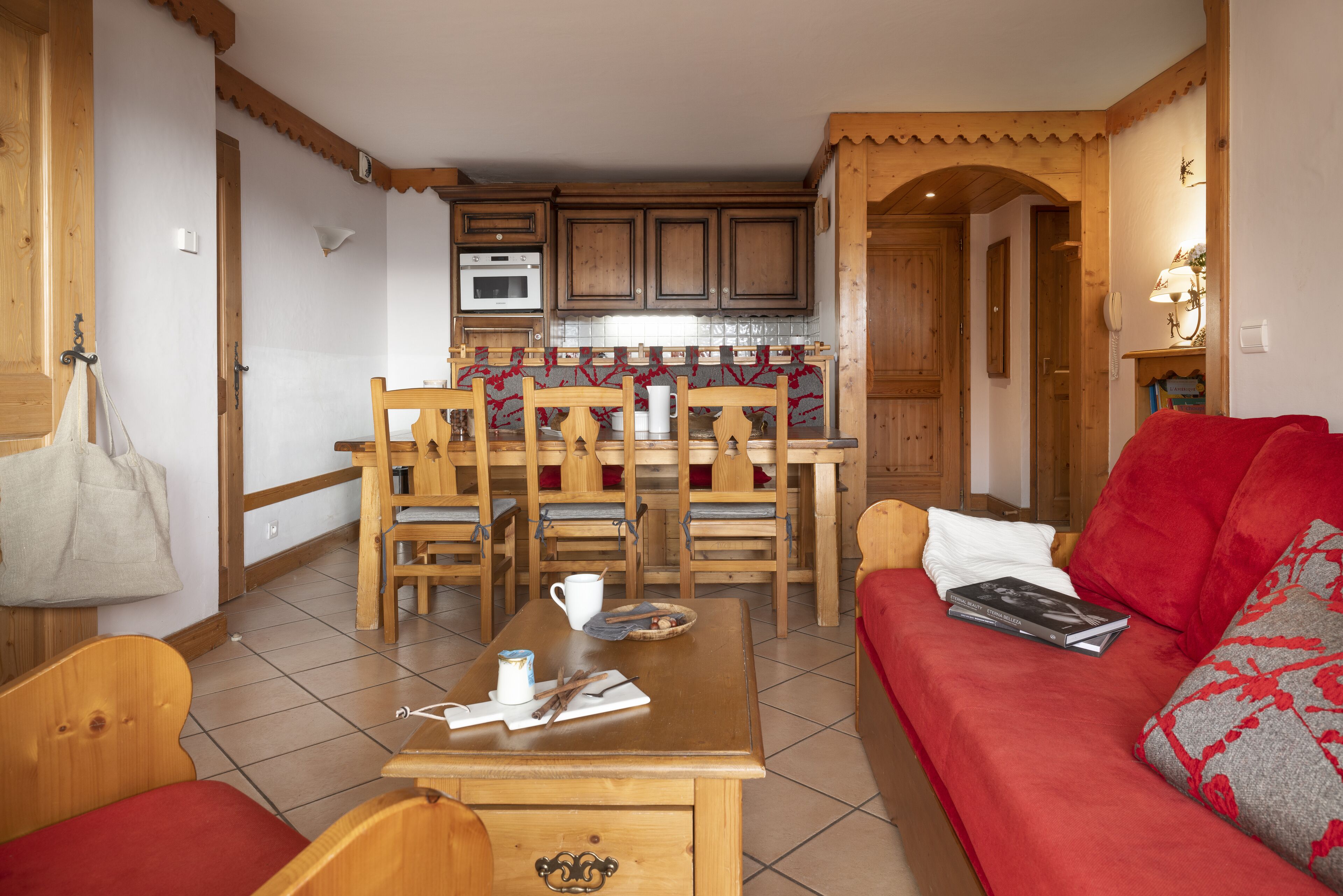 A cozy living room with a wooden table, a red sofa, and a dining area with wooden chairs and a kitchen in the background.