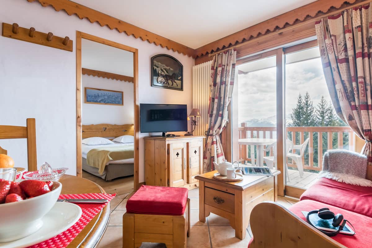 A cozy room with a bed, a TV, a wooden cabinet, a table with a bowl of fruit, and a view of the outdoors.