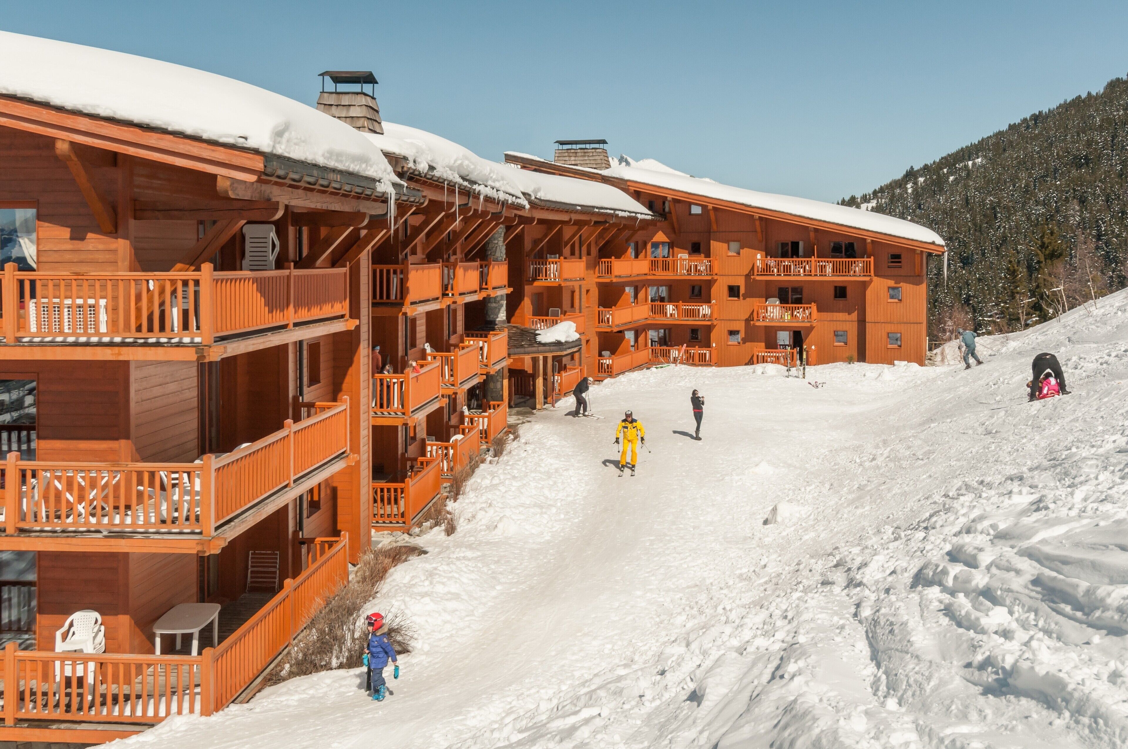 A wooden multi-story building with balconies and a ski slope in the background.