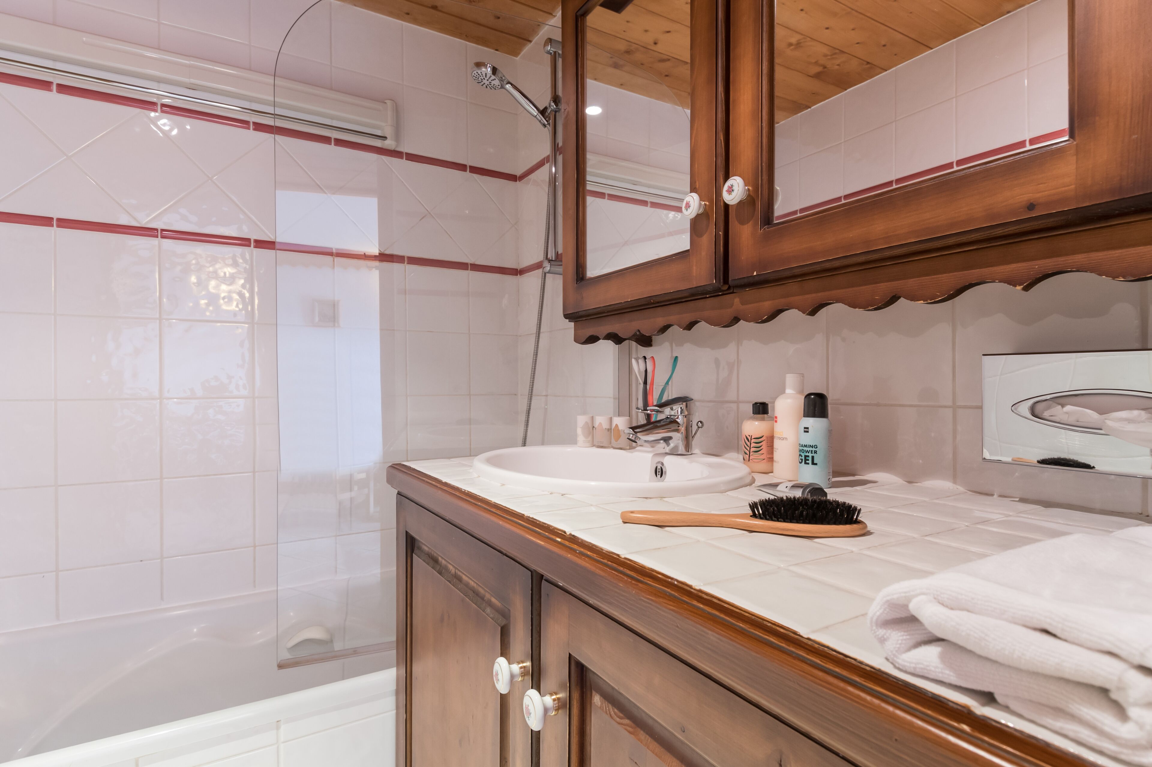 A bathroom with a wooden vanity, white tiles, a mirror, and a shower area.