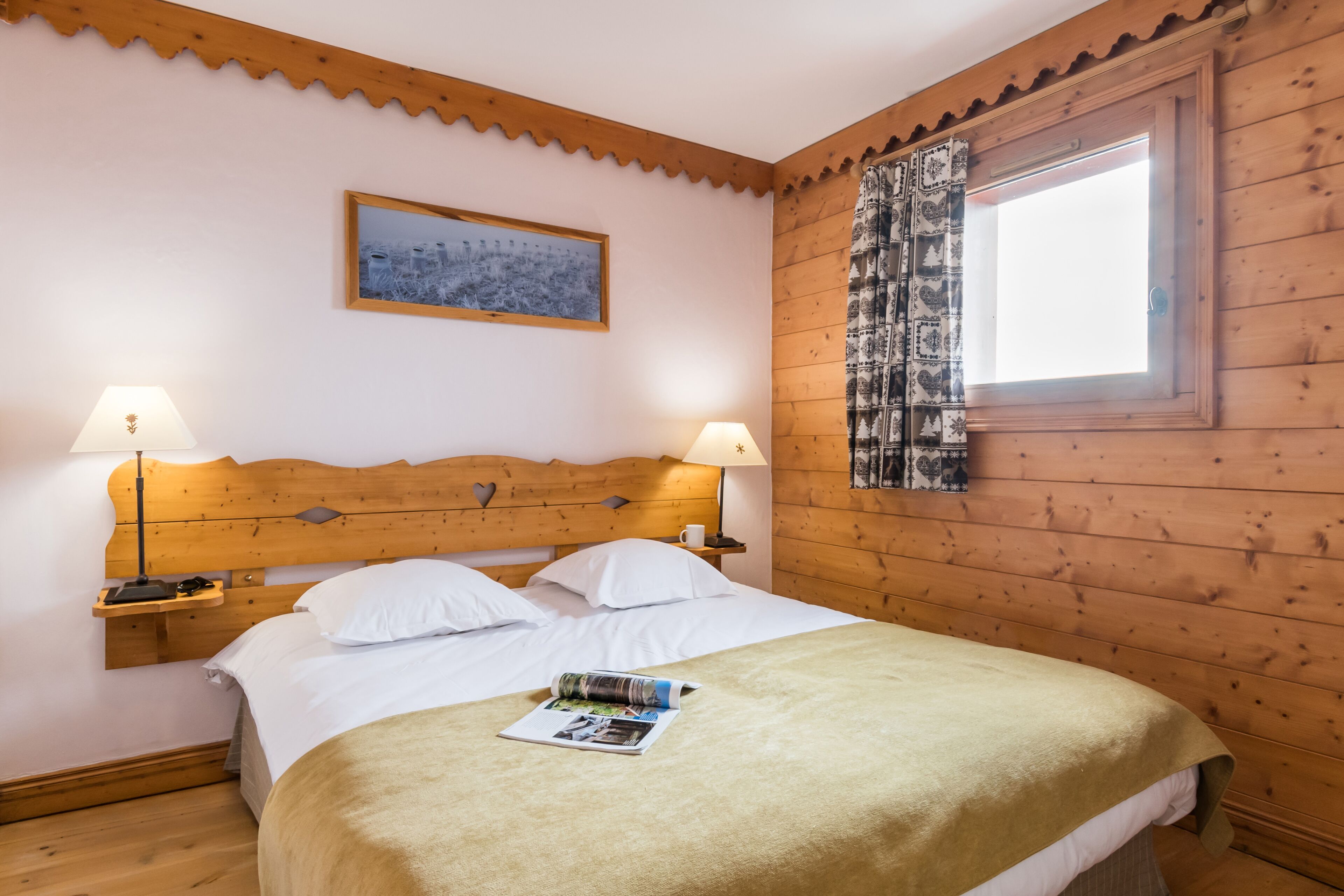 A wooden-paneled bedroom with a bed, two bedside lamps, a nightstand, a window with patterned curtains, and a framed picture on the wall.