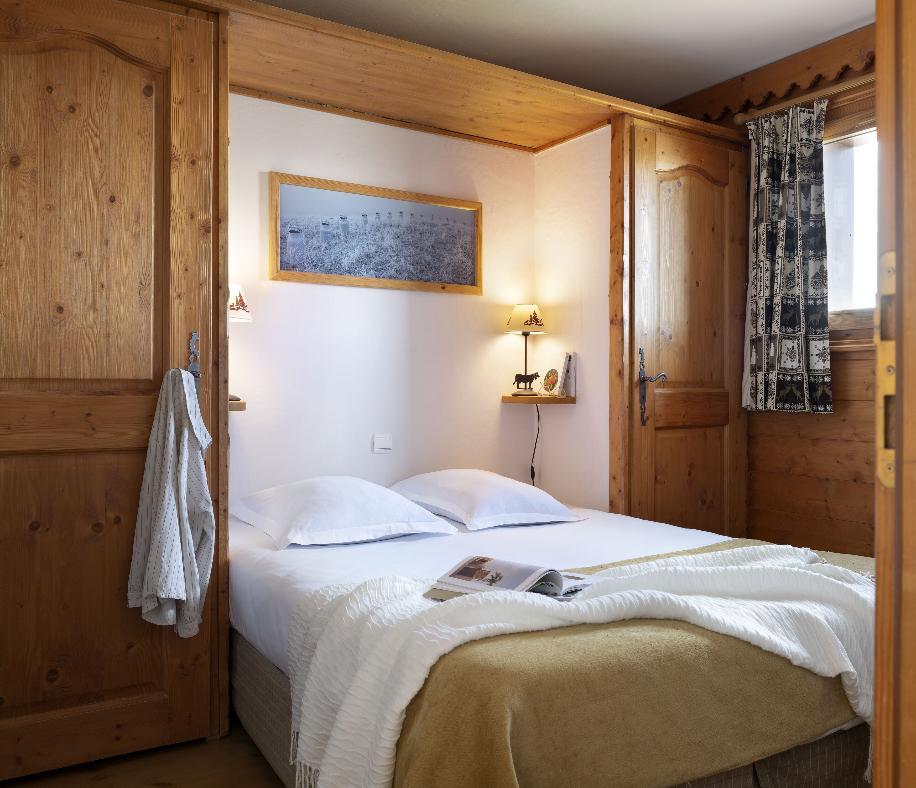 A wooden room with a bed, a hanging coat, a wall-mounted lamp, and a window with patterned curtains.