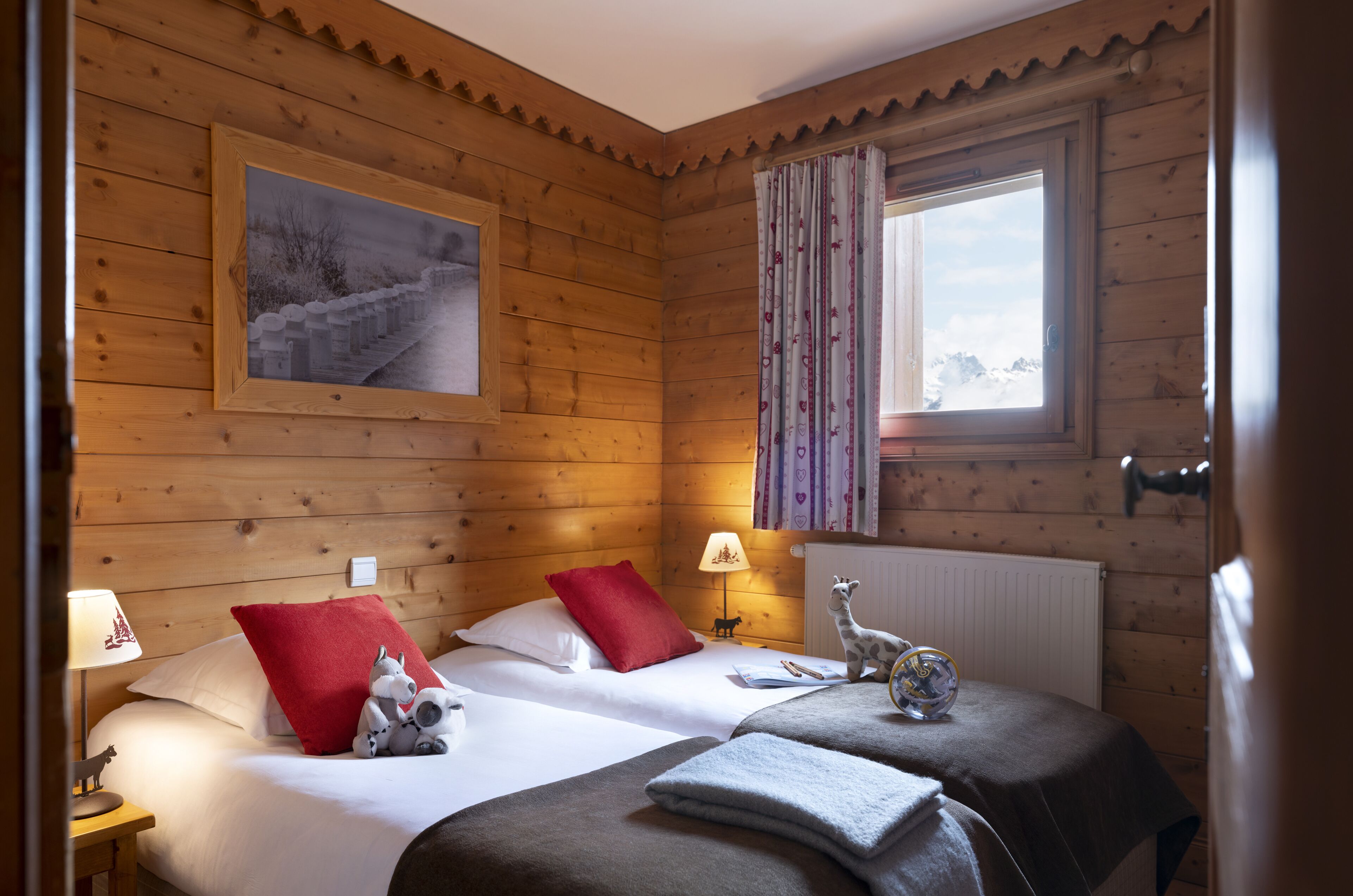 Una habitación tipo cabaña de madera con dos camas, un cuadro, una ventana con cortinas y una lámpara sobre una mesita de noche.