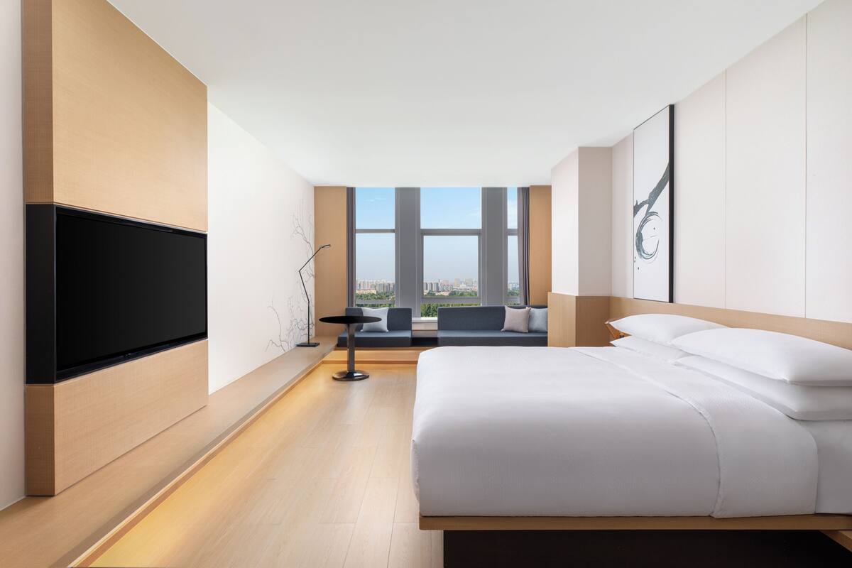 Ein modernes Hotelzimmer mit einem großen Bett, einem an der Wand montierten Flachbildfernseher, einer Sitzecke mit Sofa und Sesseln sowie einem großen Fenster mit Blick auf die Stadt.