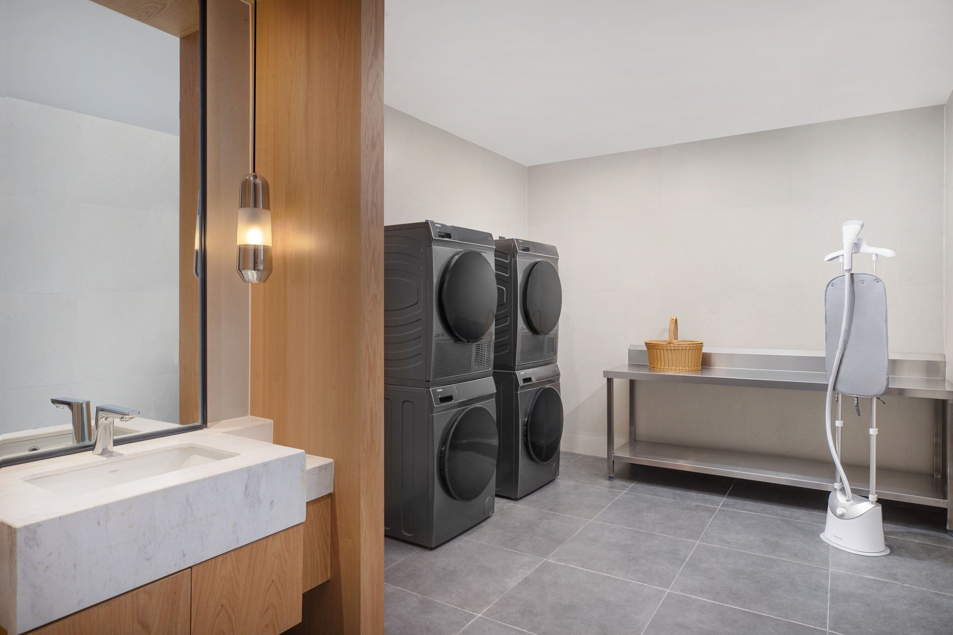 Ein modernes Badezimmer mit einem Waschbecken, zwei übereinander angeordneten Frontlader-Waschmaschinen, einem Fön und einem Korbwäscher auf einem Regal.