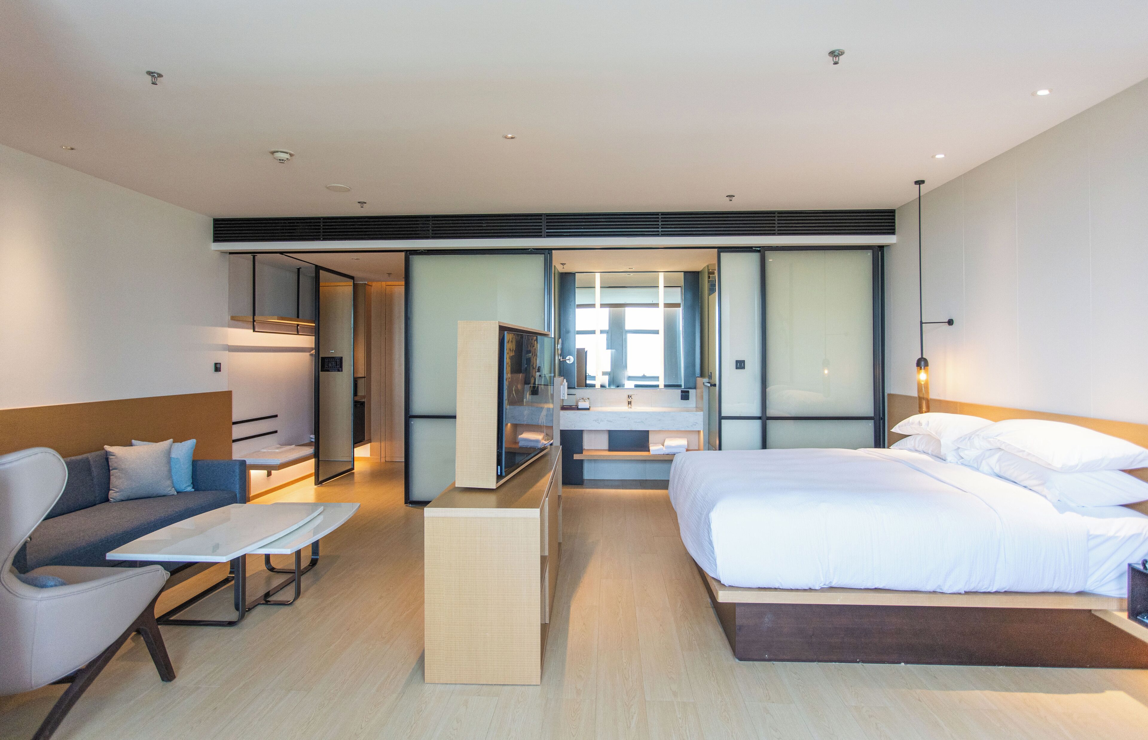 A modern hotel room with a large bed, a sofa, a small table, and a bathroom visible through an open door.