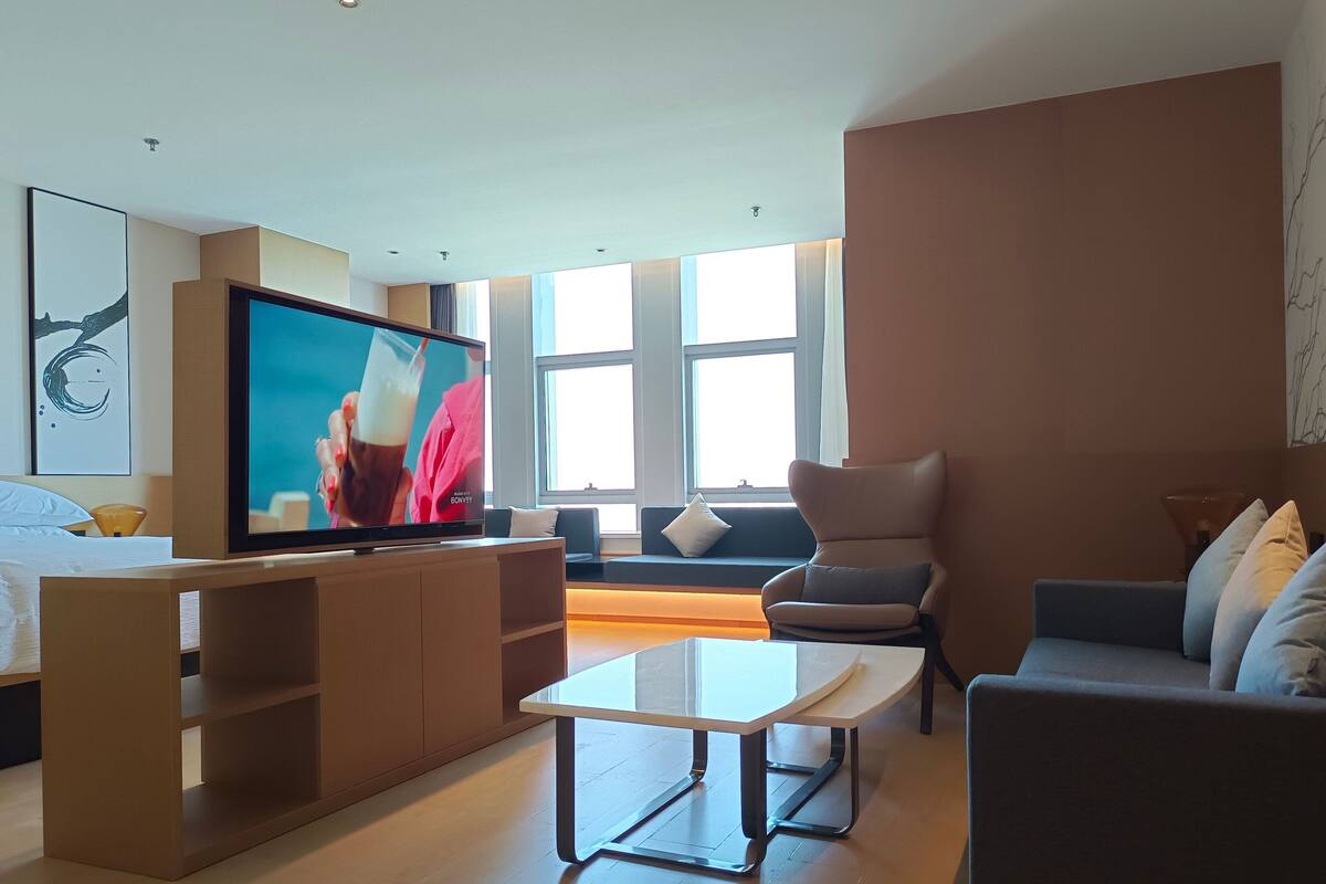 Ein modernes Wohnzimmer mit Flachbildfernseher, Sofa, Couchtisch und einem großen Fenster, das natürliches Licht hereinlässt.