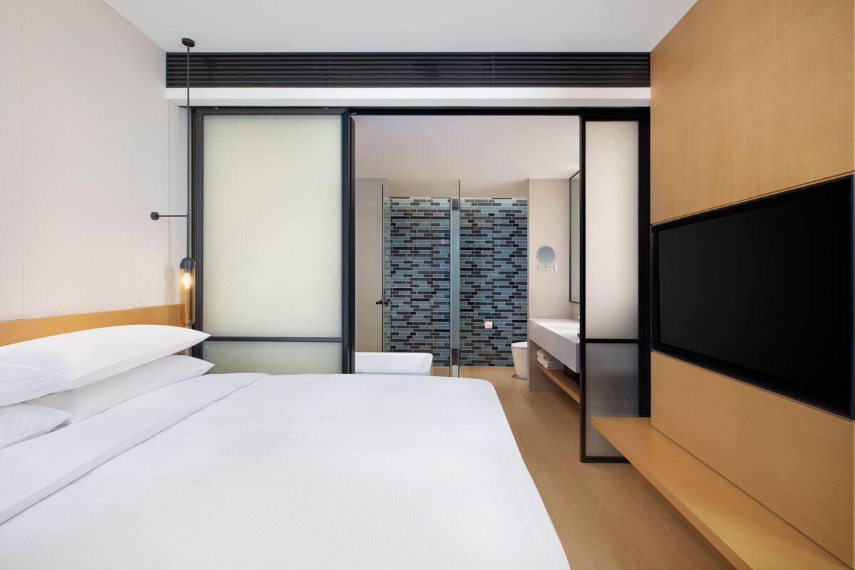Ein modernes Hotelzimmer mit einem großen Bett, einem Flachbildfernseher, einer Schiebetür zu einem Badezimmer mit Fliesen und einer Holzbank.