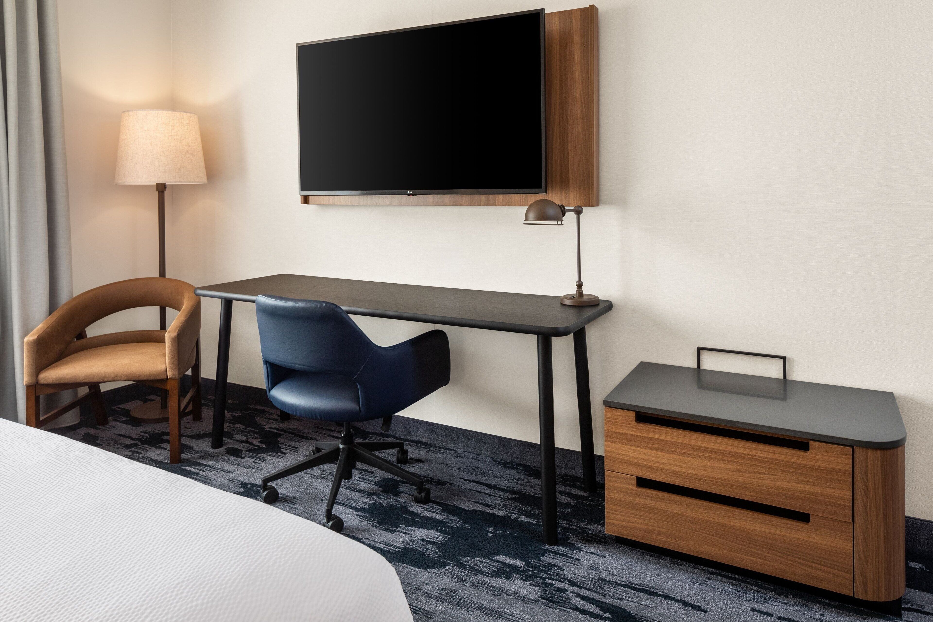 Una habitación de hotel con cama, escritorio, silla, televisor, lámpara y un mueble pequeño.